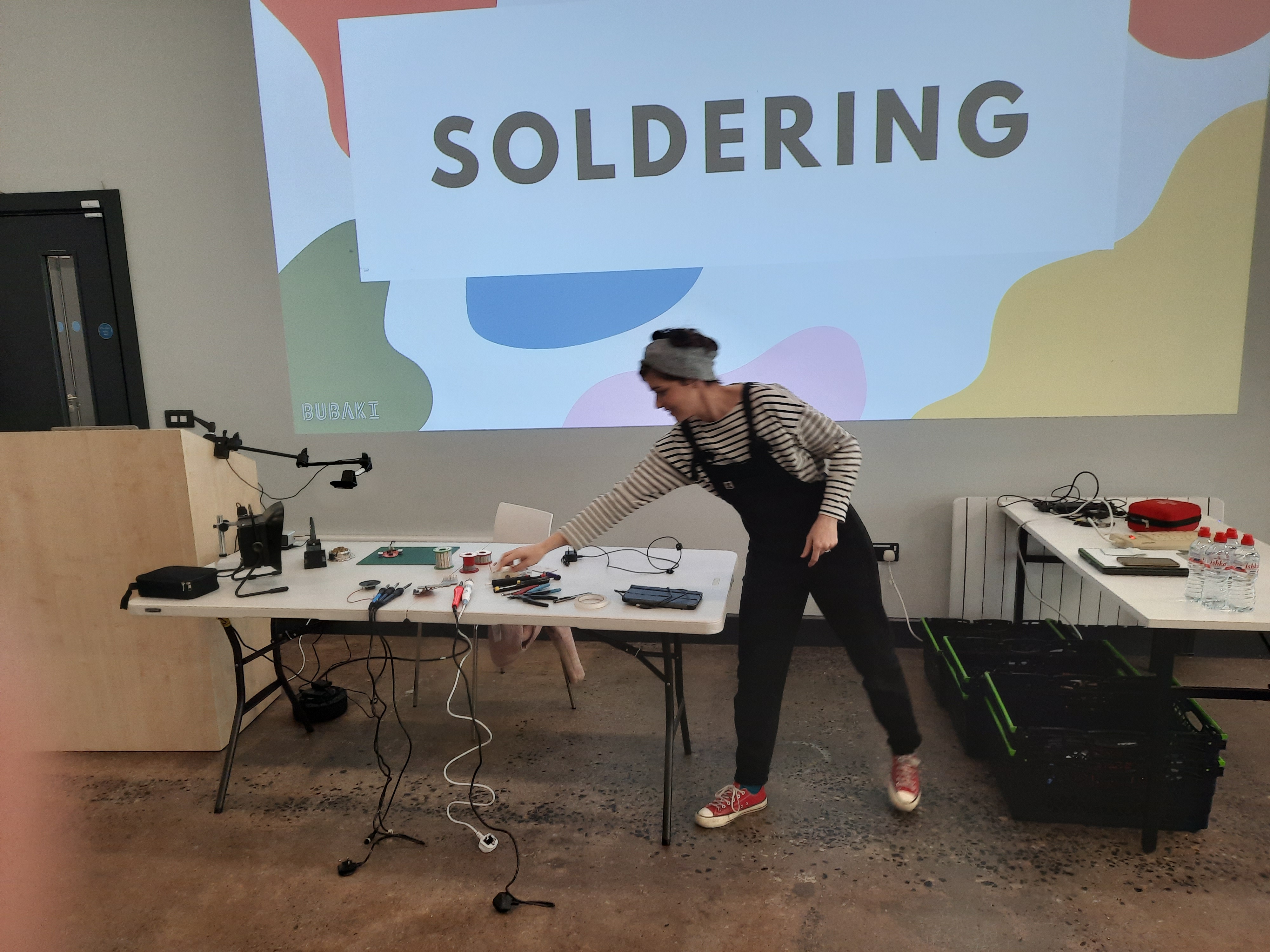 Participant soldering the board for the first time, hands and iron visible, focused expression. Workshop table.